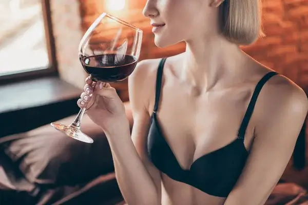 En kvinna i behå håller ett glas rött vin i handen och för det mot näsan.