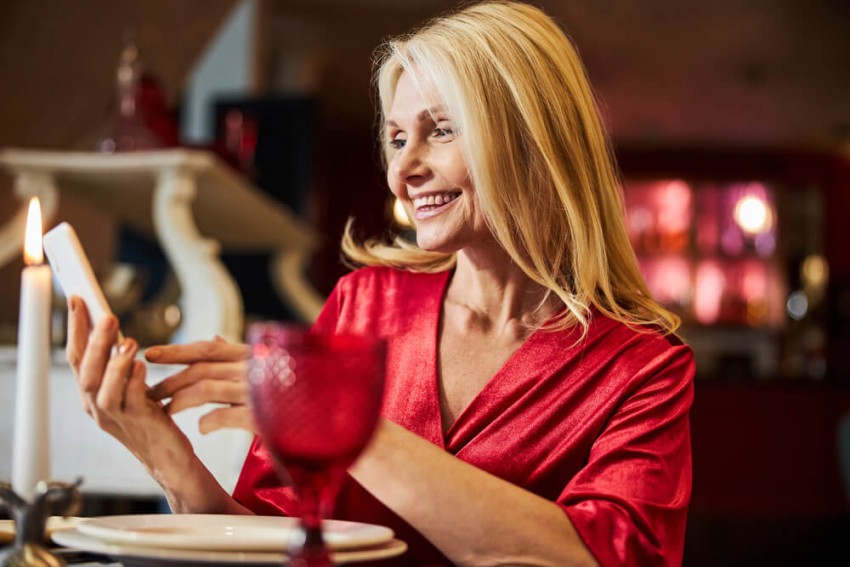 Leende mogen blond kvinna tittar på sin smartphone under en romantisk middag.